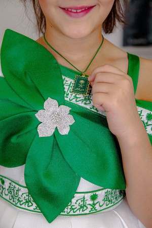 فستان بناتي لليوم الوطني لون أبيض مزين بوردة خضراء و كتابات مطرزة بدون أكمام و بكتف مكشوف و رباط في الخصر مع تعليقة بشعار سيفين و نخلة 