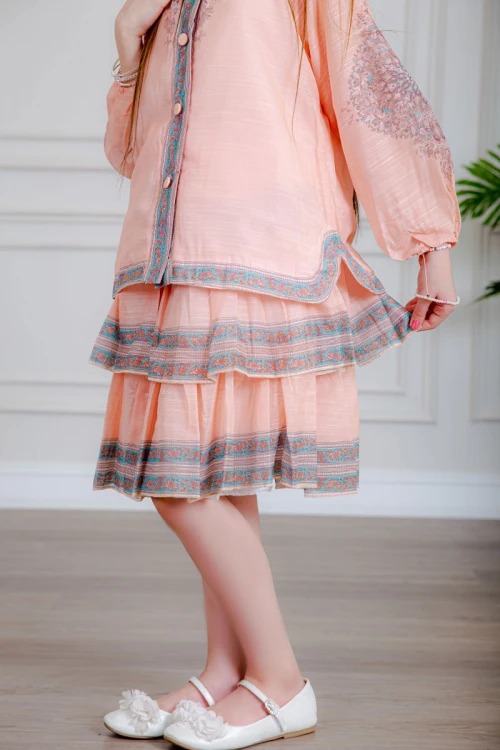 طقم تيور بناتي قطعتين لون خربزي بلوزة بياقة عالية و أزرار من الأمام ذو أكمام طويلة و تـنورة مزين بزخارف ملونة من الأطراف و الأكمام