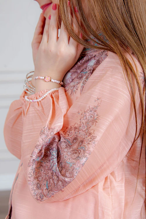 طقم تيور بناتي قطعتين لون خربزي بلوزة بياقة عالية و أزرار من الأمام ذو أكمام طويلة و تـنورة مزين بزخارف ملونة من الأطراف و الأكمام