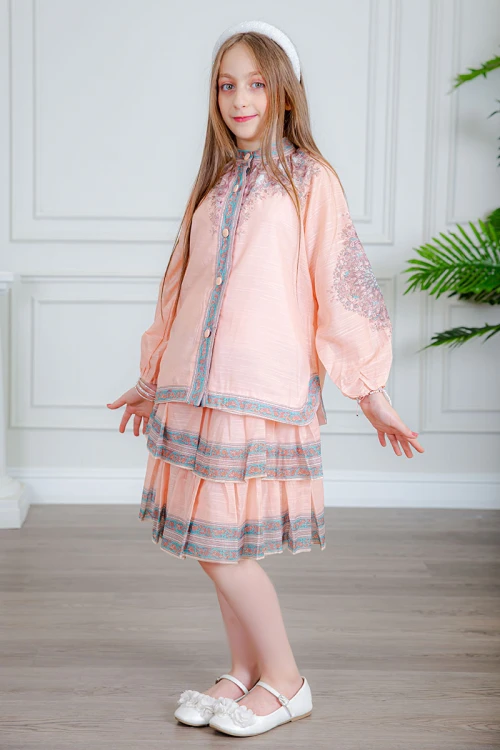 طقم تيور بناتي قطعتين لون خربزي بلوزة بياقة عالية و أزرار من الأمام ذو أكمام طويلة و تـنورة مزين بزخارف ملونة من الأطراف و الأكمام