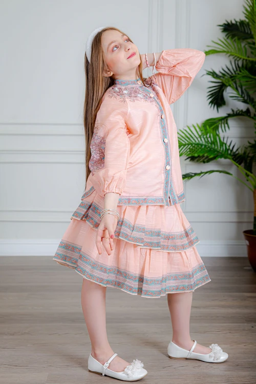 طقم تيور بناتي قطعتين لون خربزي بلوزة بياقة عالية و أزرار من الأمام ذو أكمام طويلة و تـنورة مزين بزخارف ملونة من الأطراف و الأكمام