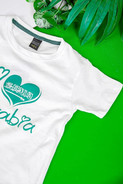 تيشيرت اليوم الوطني السعودي لون أبيض مزين بكتابات و رسومات من الأمام باللون الأخضر