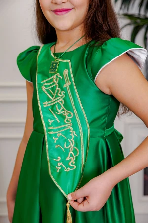 فستان بناتي لليوم الوطني لون أخضر مزين بوشاح من الأمام بتطريز عبارة المملكة العربية السعودية باللون الذهبي بأكمام قصيرة و رباط في الخلف مع تعليقة بشعار سيفين و نخلة 