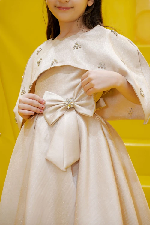 فستان بناتي لون بيج ياقة دائرية بدون أكمام مع فيونكة من الأمام و رباط في الخصر و بكاب علوي منفصل على الكتف مزين بفصوص الكريستال
