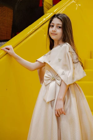 فستان بناتي لون بيج ياقة دائرية بدون أكمام مع فيونكة من الأمام و رباط في الخصر و بكاب علوي منفصل على الكتف مزين بفصوص الكريستال  