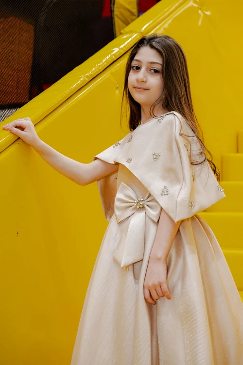 فستان بناتي لون بيج ياقة دائرية بدون أكمام مع فيونكة من الأمام و رباط في الخصر و بكاب علوي منفصل على الكتف مزين بفصوص الكريستال