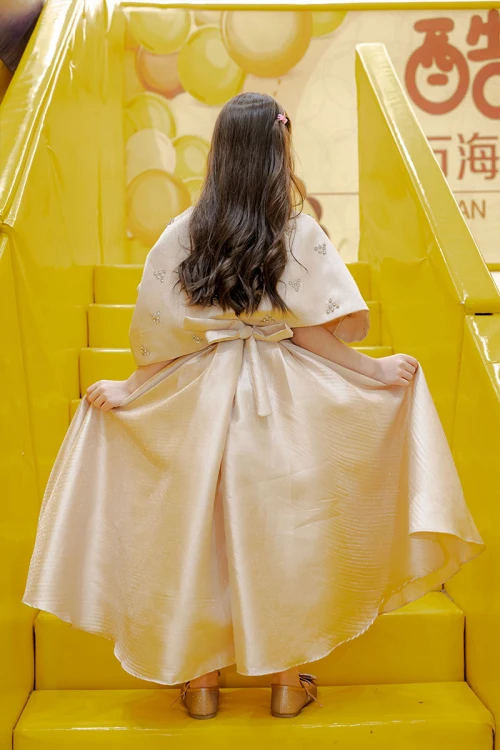 فستان بناتي لون بيج ياقة دائرية بدون أكمام مع فيونكة من الأمام و رباط في الخصر و بكاب علوي منفصل على الكتف مزين بفصوص الكريستال